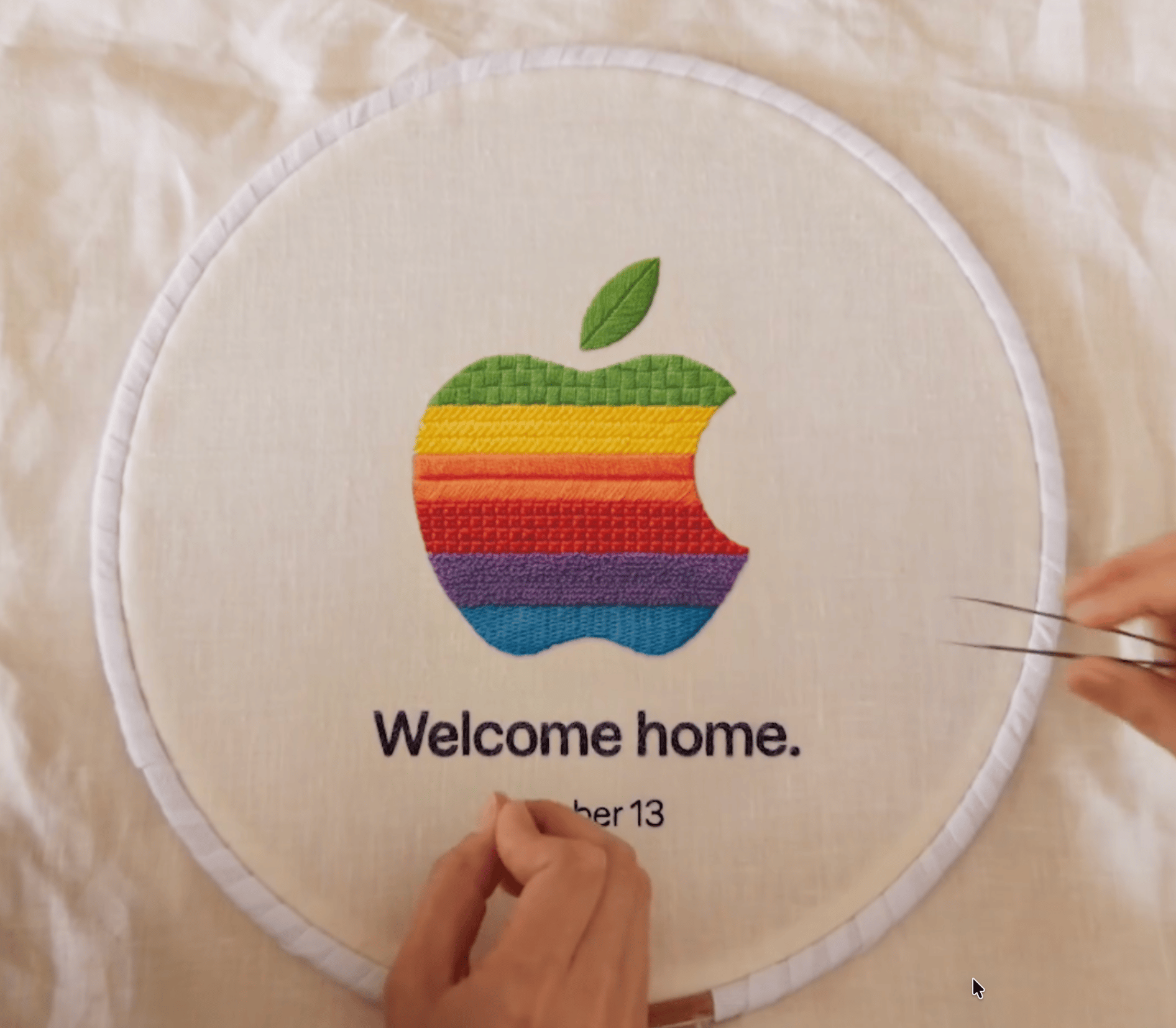 Rainbow-striped apple logo embroidered on cream fabric in a hoop, with 'Welcome home.' text and hands holding tweezers.