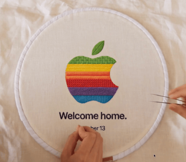 Rainbow-striped apple logo embroidered on cream fabric in a hoop, with 'Welcome home.' text and hands holding tweezers.