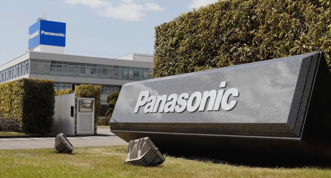 Panasonic sign on grass in front of a modern office building topped with a blue Panasonic logo, framed by green hedges
