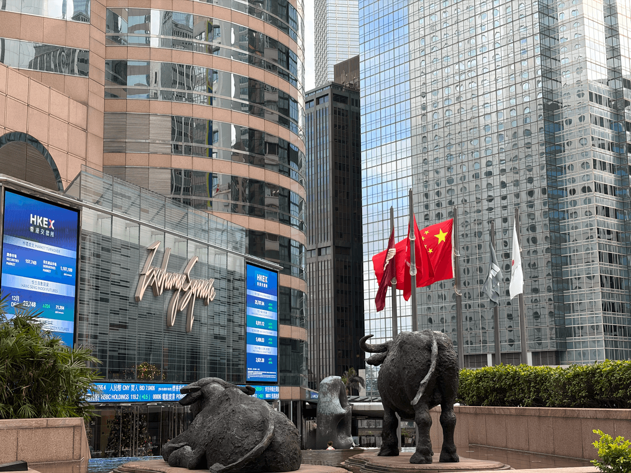 Exterior view of Hong Kong Exchanges and Clearing’s Exchange Square building in Central, with stock market ticker screens, Chinese and Hong Kong flags, and the bronze bull and bear statues in the foreground.