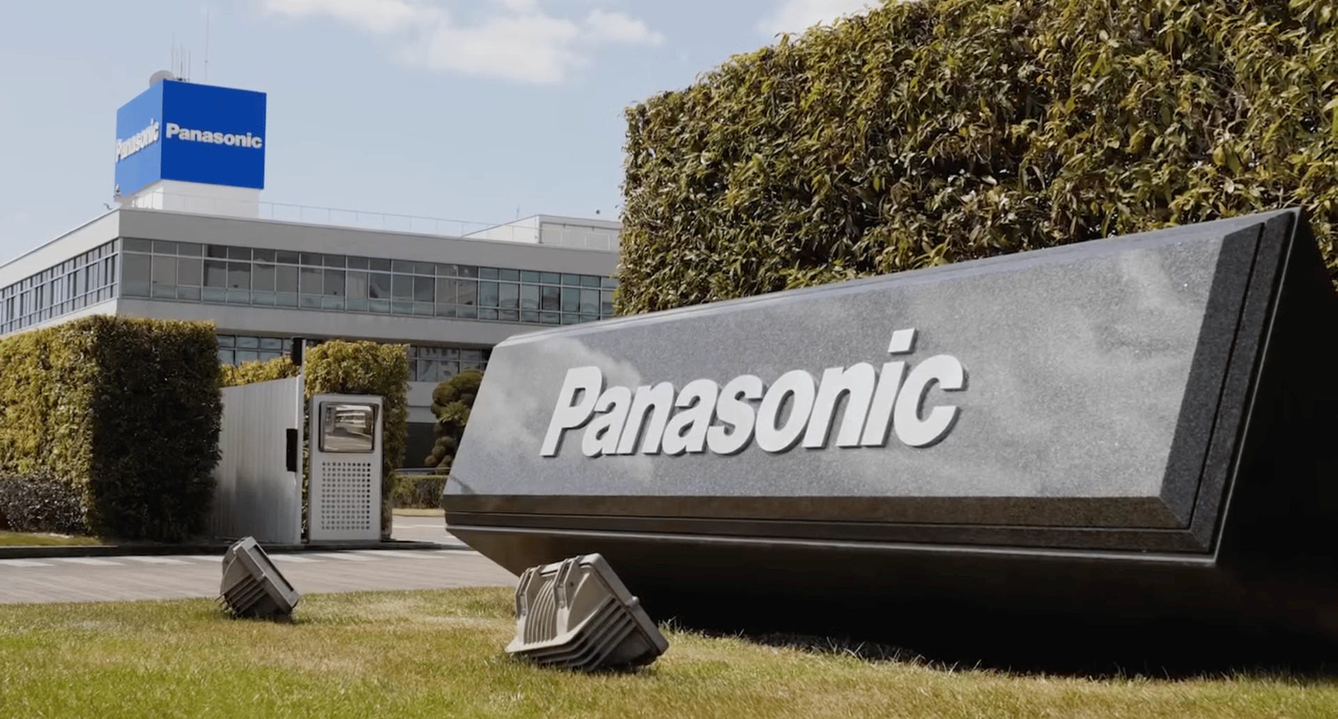 Panasonic sign on grass in front of a modern office building topped with a blue Panasonic logo, framed by green hedges